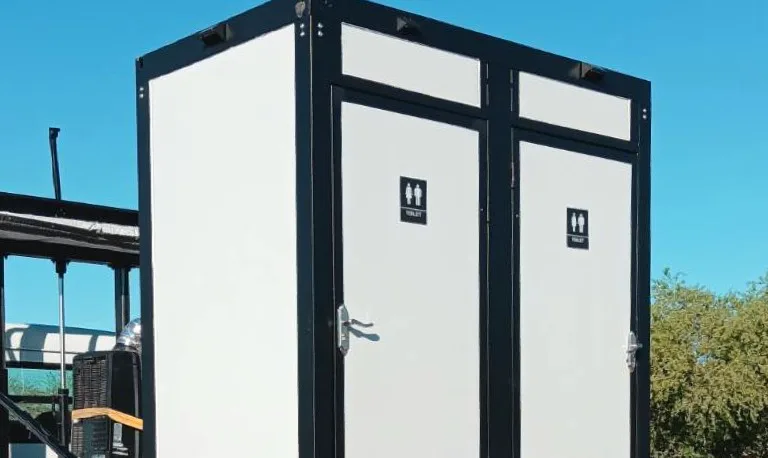 "Portable restroom trailer with two doors and black trim on site under clear blue sky."